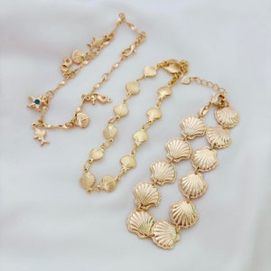May include: Two gold necklaces with a seashell design. The longer necklace has a larger seashell pendant and the shorter necklace has smaller seashells.
