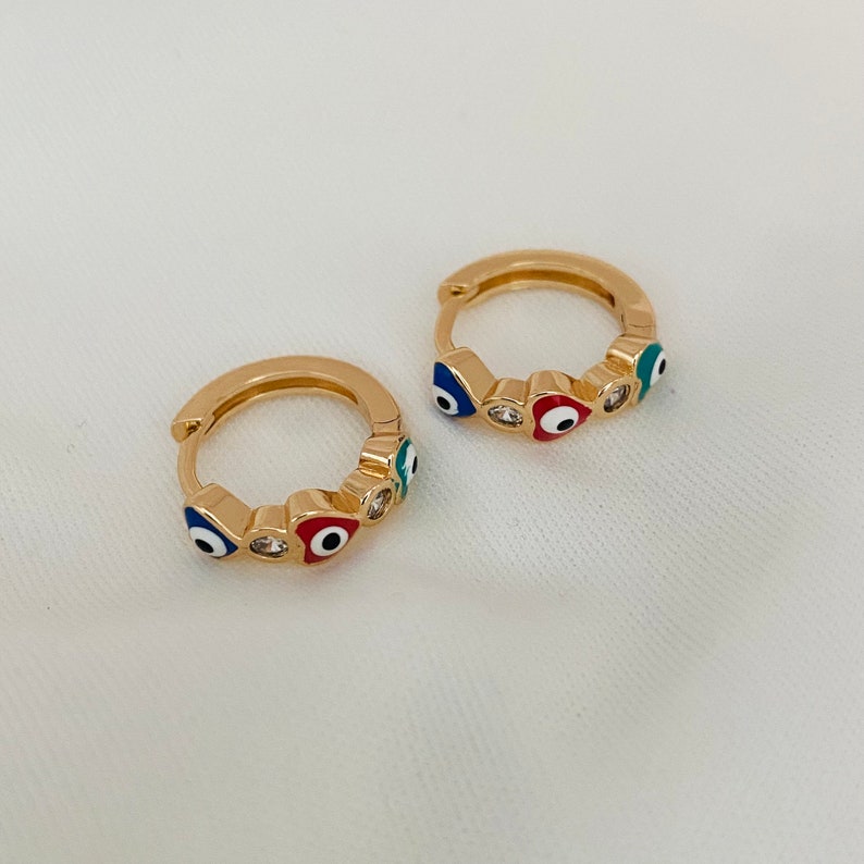 K&ouml;nnte beinhalten: Ein Paar goldene Creolen mit einem Design aus blauen, roten und gr&uuml;nen b&ouml;sen Augen. Die Ohrringe sind klein und zart.