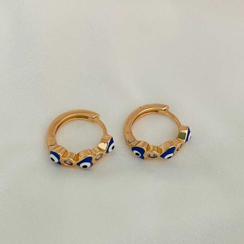 K&ouml;nnte beinhalten: Ein Paar goldene Creolen mit blauen b&ouml;sen Augen-Anh&auml;ngern und klaren Strasssteinen.