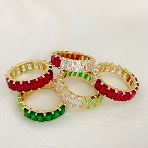May include: Four gold rings with colorful gemstones. The rings are stacked on top of each other. The top ring has clear gemstones, the second ring has green gemstones, the third ring has red gemstones, and the bottom ring has red gemstones.