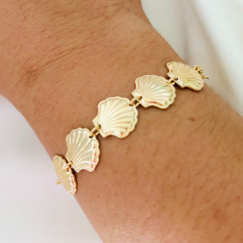 May include: A gold bracelet with a chain and five shell-shaped charms. The bracelet is on a person's wrist.