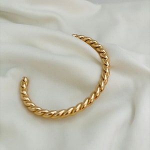 Könnte beinhalten: Ein goldfarbenes, gedrehtes Armreif-Armband. Das Armband besteht aus einem dicken, seilartigen Metall.