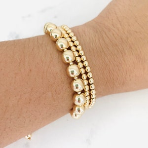 May include: Three gold beaded bracelets stacked on a wrist. The bracelets are made of round, shiny gold beads.
