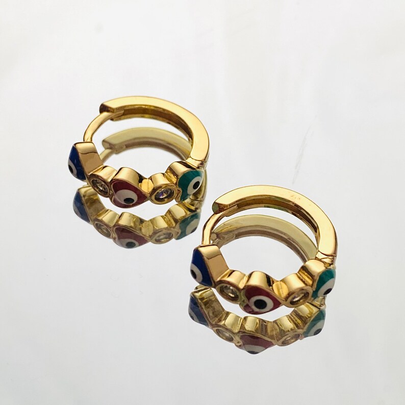 K&ouml;nnte beinhalten: Goldene Creolen mit einem Design aus mehreren blauen, roten und gr&uuml;nen b&ouml;sen Augen. Jedes Auge hat eine wei&szlig;e Mitte und ist von einem goldenen Rand umgeben. Zwischen den Augen befinden sich auch kleine klare Strasssteine.