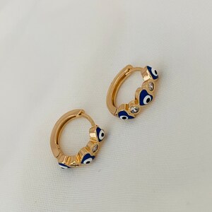 K&ouml;nnte beinhalten: Ein Paar goldene Creolen mit blauen und wei&szlig;en b&ouml;sen Augen-Anh&auml;ngern. Die Ohrringe sind aus Metall und haben eine gl&auml;nzende Oberfl&auml;che.
