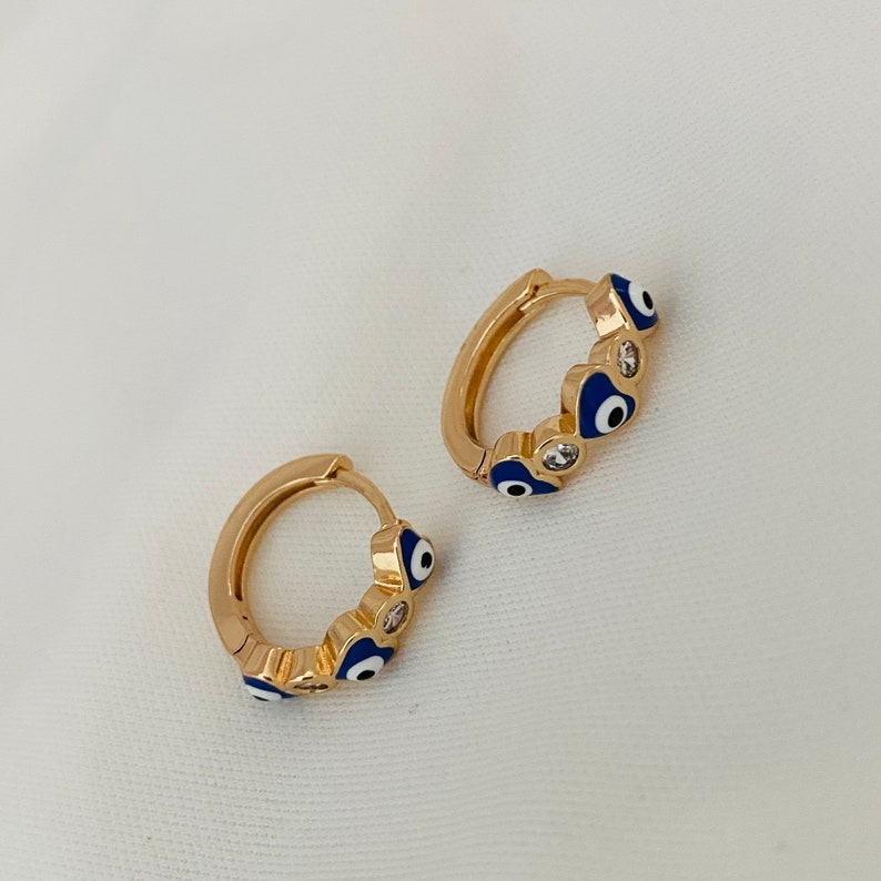 K&ouml;nnte beinhalten: Ein Paar goldene Creolen mit blauen und wei&szlig;en b&ouml;sen Augen-Anh&auml;ngern. Die Ohrringe sind aus Metall und haben eine gl&auml;nzende Oberfl&auml;che.