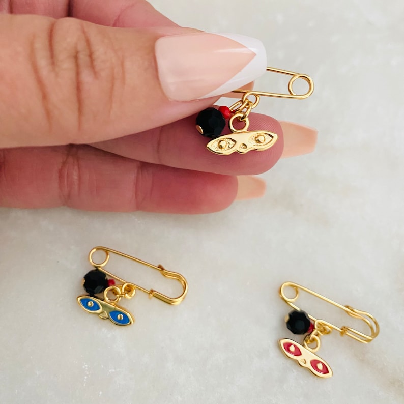 May include: Three gold safety pin brooches with black beads and a small gold mask charm. The masks have red, blue, and black eyes.