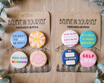 Può includere: Due set di pulsanti motivazionali con messaggi ispiratori. Il primo set include "Va bene non stare bene", "Sii gentile", "Sei abbastanza", e "La salute mentale conta". Il secondo set include "Rimani in vita", "Va bene commettere errori", "Sii gentile con la tua mente", e "Amore per se stessi".