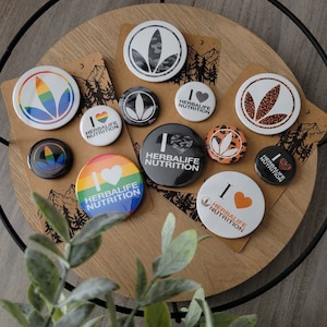 May include: A collection of round, colorful buttons with the text "I ♥ Herbalife Nutrition" on them. The buttons feature various designs, including a rainbow, a camouflage pattern, and a leopard print.
