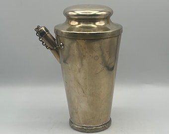 Puede incluir: Una coctelera de plata con una cadena dorada y un pico. La coctelera tiene una banda decorativa alrededor de la parte inferior.