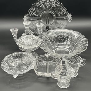 May include: A collection of clear glass serving dishes with etched floral designs. The set includes a large serving platter, a bowl, a divided dish, a creamer, a sugar bowl, a small bowl, and a candle holder.