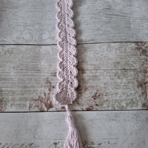 May include: A light pink, handmade crochet bookmark. The bookmark has a scalloped edge and a tassel. It's made of soft yarn, ideal for marking pages in books. The bookmark is a delicate craft item.