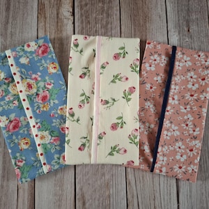 May include: Three rectangular fabric pouches featuring floral patterns. The left pouch is blue with pink and yellow roses and a white trim with red dots. The centre is cream with pink rosebuds and pink trim. The right is pink with white flowers and a dark blue trim.