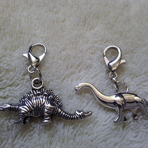 May include: Two silver dinosaur charms with lobster claw clasps. One charm depicts a Stegosaurus with detailed ridges, and the other is a long-necked Brontosaurus. These charms are suitable for jewellery making or as decorative accents.