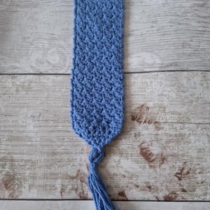 May include: A handcrafted, blue crochet bookmark with a textured design. The bookmark is rectangular, featuring a small hole at the top and a tassel at the bottom. The tassel is made from the same blue yarn as the bookmark.