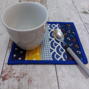 May include: A blue and white fabric coaster with a yellow, blue, and white checked pattern. A white ceramic mug and a silver spoon are on top of the coaster.