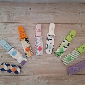 May include: A collection of seven fabric dummy clips featuring various patterns and colours, including geometric shapes, stripes, and floral prints. Each clip has a plastic fastener at the top and a matching snap button closure on the fabric strap.