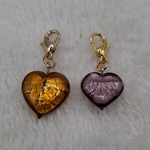 May include: Two gold heart-shaped charms with a gold clasp. The charm on the left is amber with gold flecks. The charm on the right is purple with gold flecks.