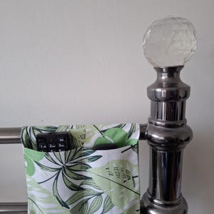 May include: A fabric bed tidy with a tropical leaf design in green and white, attached to a metal bed frame. A black remote control is visible in one of the pockets. The bed frame has a decorative, crystal-like ball on top.