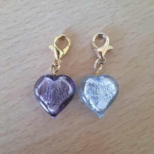 May include: Two heart-shaped charms with gold-coloured clasps. One charm is a translucent purple with a textured interior, and the other is a translucent blue with a silver textured interior. The charms are likely for jewellery or crafts.