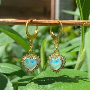 May include: A pair of gold-coloured hoop earrings. Each earring has a heart-shaped opal pendant with a halo of small, clear crystals. The opals show a mix of blue and green hues. The earrings are hanging on a wooden stick against a backdrop of green foliage.