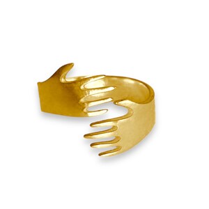 Peut inclure: Une bague dorée en forme de deux mains qui s'étreignent. La bague a une finition lisse et polie et est conçue pour s'enrouler autour du doigt. Les mains sont détaillées avec des doigts individuels, créant un bijou unique et symbolique.
