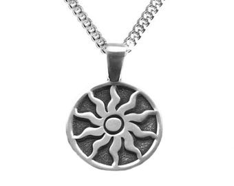 May include: A silver-toned metal pendant necklace featuring a circular charm with a raised sun design. The sun has a central circular core surrounded by twelve wavy, flame-like rays. The pendant hangs from a matching silver-toned curb chain.