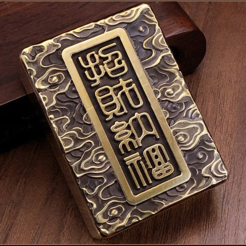 May include: A rectangular, antique-style lighter with a brass finish. The lighter features an embossed cloud pattern and a rectangular brass plate with Chinese characters. The overall design is detailed and ornate.