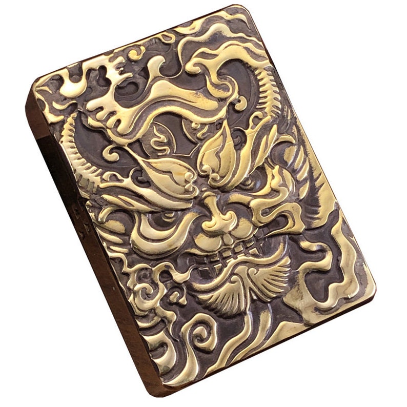 May include: A rectangular, brass-colored metal object with a detailed relief of a mythical creature's face. The design features intricate patterns and textures, with a dark background and gold-colored raised elements. The object has a dark brown border.