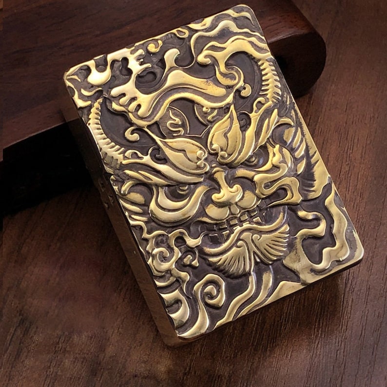 May include: A rectangular, brass-colored lighter with an intricate, embossed design. The design features a detailed, stylized face with swirling patterns and ornate details. The lighter has a dark, contrasting background, highlighting the raised relief.