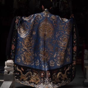 May include: A person wearing a blue and gold embroidered robe. The robe features intricate dragon and floral patterns. The bottom hem is black with gold dragon designs. The person is standing in a dimly lit room.