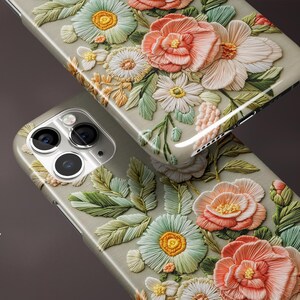 K&ouml;nnte beinhalten: Zwei Handyh&uuml;llen mit gl&auml;nzendem Finish, mit einem 3D-Blumendesign. Die H&uuml;llen zeigen eine Vielzahl von Blumen in Pfirsich-, Wei&szlig;- und Gr&uuml;nt&ouml;nen mit aufwendigen Details. Die H&uuml;llen sind hellgrau.
