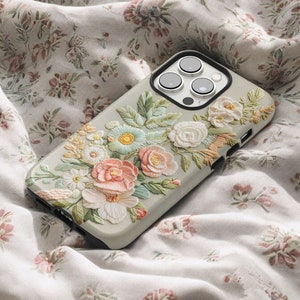 iPhone case with an intricate floral embroidery. lying on top of a soft fabric background with delicate flowers and leaves. The case features focus stacking, cottagecore aesthetics, vintage style