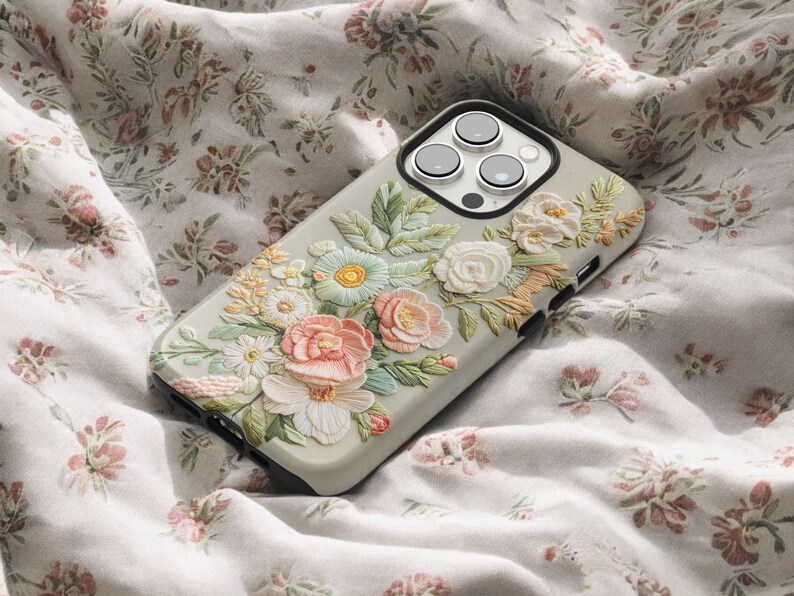 iPhone case with an intricate floral embroidery. lying on top of a soft fabric background with delicate flowers and leaves. The case features focus stacking, cottagecore aesthetics, vintage style