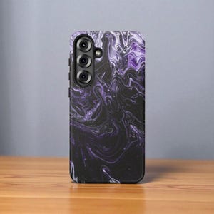 May include: A rectangular phone case with a swirling, marbled design in shades of black, white, and purple. The case has a camera module with three lenses. The case is resting on a wooden surface.