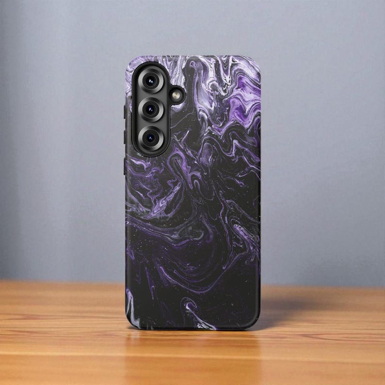 May include: A rectangular phone case with a swirling, marbled design in shades of black, white, and purple. The case has a camera module with three lenses. The case is resting on a wooden surface.