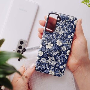 May include: A navy blue phone case with a floral pattern in white and light blue is held in a person's hand. The case is next to a white phone and a white face mask. Green plants are in the background.