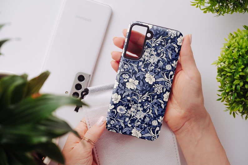 May include: A navy blue phone case with a floral pattern in white and light blue is held in a person's hand. The case is next to a white phone and a white face mask. Green plants are in the background.