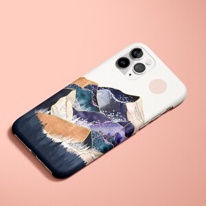 May include: White iPhone case with a mountain range design. The case features a navy blue, tan, teal, and purple mountain landscape with gold accents. The phone case is on a pink background.