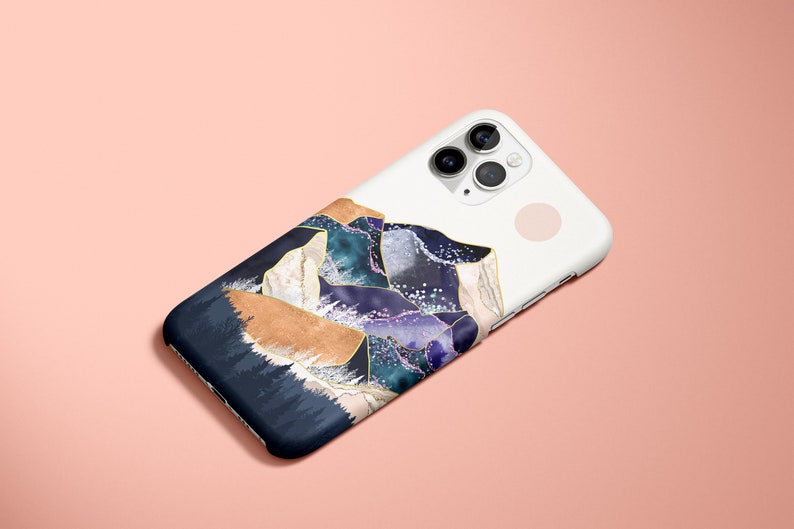 May include: White iPhone case with a mountain range design. The case features a navy blue, tan, teal, and purple mountain landscape with gold accents. The phone case is on a pink background.