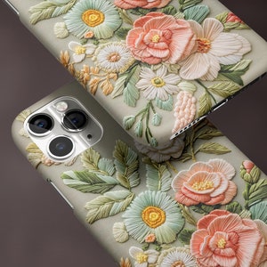 K&ouml;nnte beinhalten: Zwei Handyh&uuml;llen mit matter Oberfl&auml;che, verziert mit detaillierter Blumenstickerei in Pastellt&ouml;nen. Das Design umfasst pfirsichfarbene, wei&szlig;e und gr&uuml;ne Blumen mit aufwendigen Bl&auml;ttermustern. Der Text "MATTE FINISH" ist sichtbar.