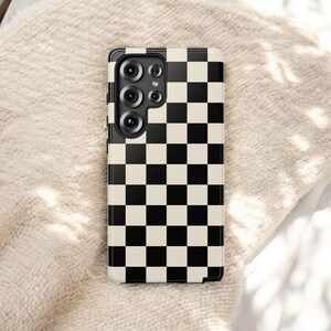 May include: A black and cream checkered phone case. The case has a glossy finish and features a grid pattern of alternating black and cream squares. The phone case has a camera module with multiple lenses.
