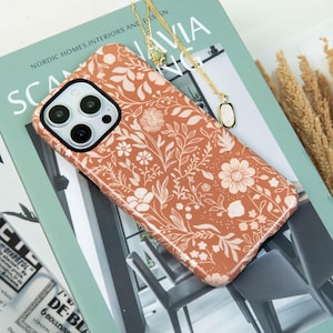 Op de afbeelding: Een terracotta telefoonhoesje met een wit bloemmotief, liggend op een boek. Een gouden ketting met een hanger ligt over de hoes. De titel van het boek luidt "NORDIC HOMES. INTERIORS AND DESIGN".