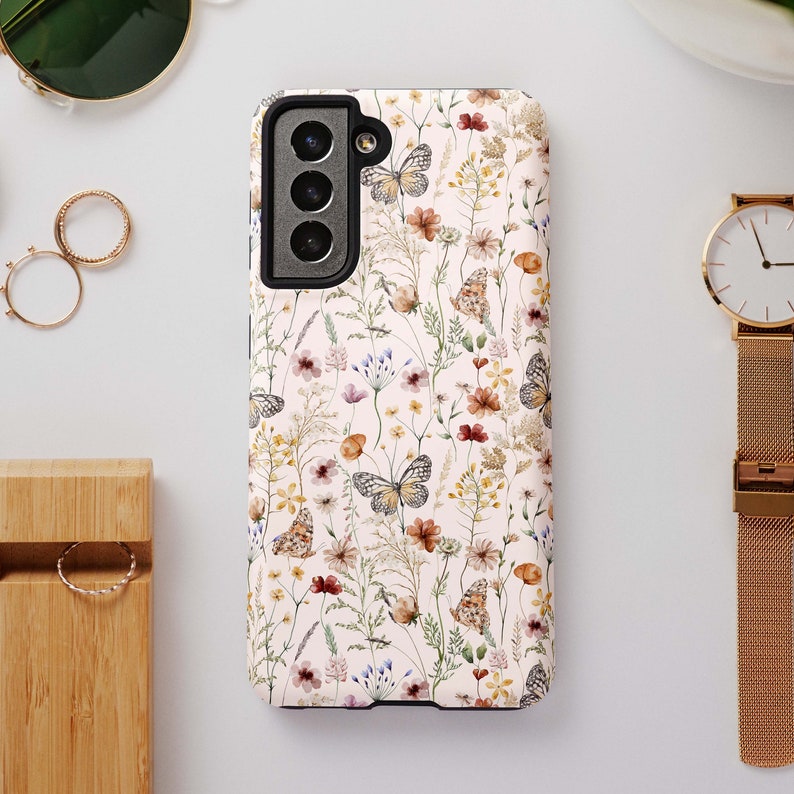 May include: A white phone case with a floral pattern featuring butterflies and wildflowers. The case has a black camera cutout and a black border.
