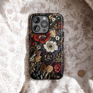 K&ouml;nnte beinhalten: Schwarze Smartphone-H&uuml;lle mit floralem Design. Die H&uuml;lle hat einen dunklen Hintergrund mit einer Vielzahl farbenfroher Blumen, darunter rote, wei&szlig;e, lila und gelbe Bl&uuml;ten. Sie liegt auf einem wei&szlig;en Stoff mit floralem Muster.