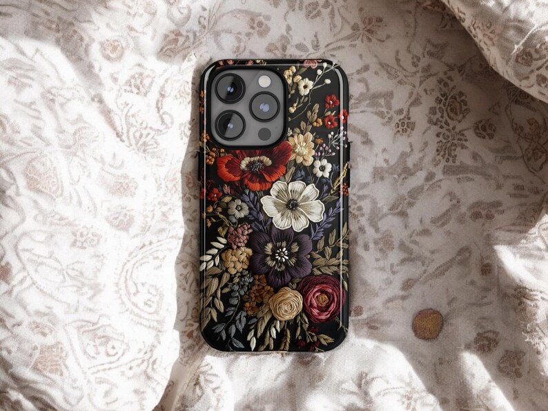 K&ouml;nnte beinhalten: Schwarze Smartphone-H&uuml;lle mit floralem Design. Die H&uuml;lle hat einen dunklen Hintergrund mit einer Vielzahl farbenfroher Blumen, darunter rote, wei&szlig;e, lila und gelbe Bl&uuml;ten. Sie liegt auf einem wei&szlig;en Stoff mit floralem Muster.