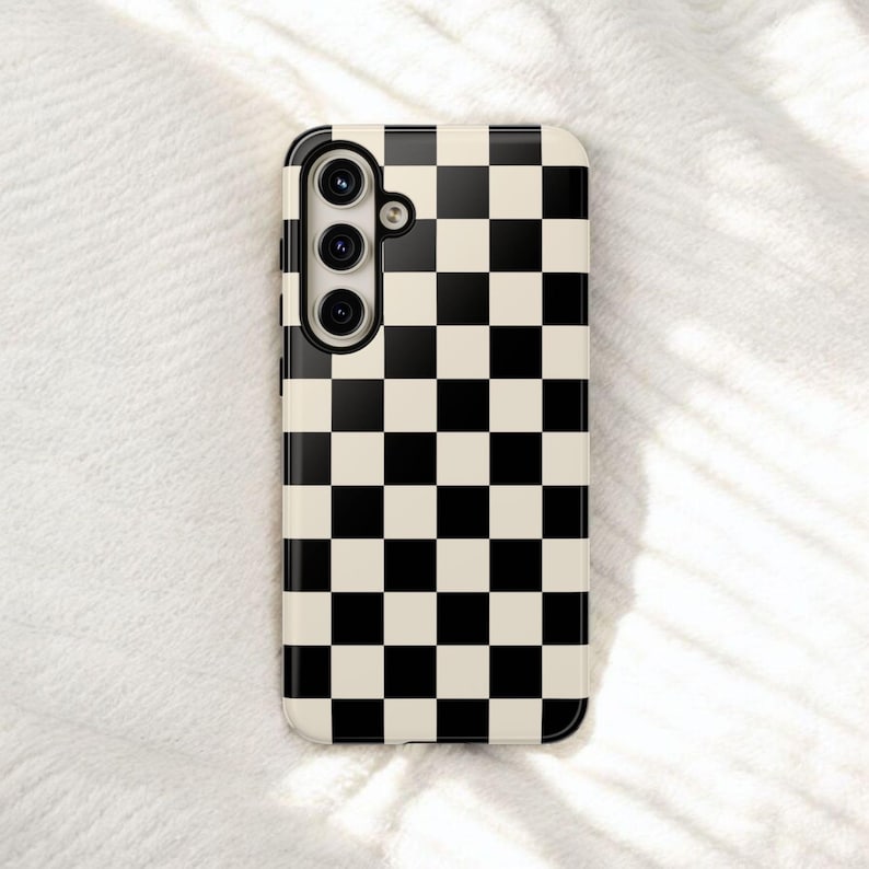 May include: A phone case with a black and cream checkerboard pattern. The case has a camera module with three lenses and a flash. The case is rectangular with rounded corners.