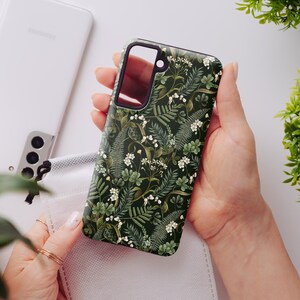 May include: A green phone case with a floral pattern featuring white flowers and green leaves. The case has a black border and a clear camera cutout.