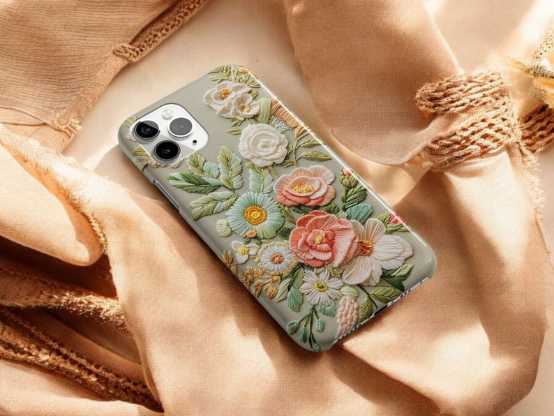 iPhone case with an intricate floral embroidery. lying on top of a soft fabric background with delicate flowers and leaves. The case features focus stacking, cottagecore aesthetics, vintage style