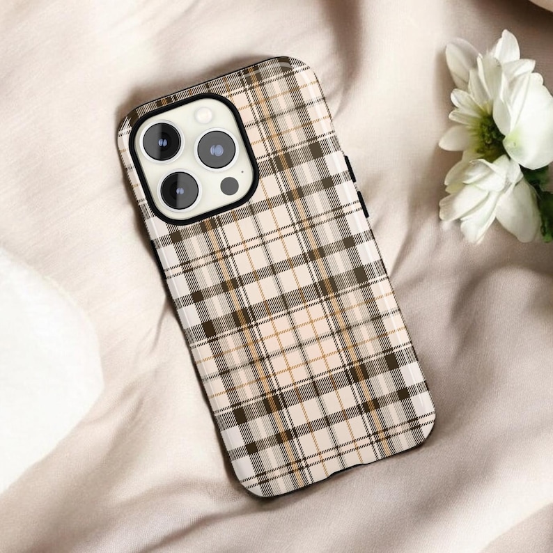 May include: A phone case with a plaid pattern in shades of brown, tan, and white. The case has a black border and a camera cutout. The case is lying on a beige surface, with white flowers in the background.
